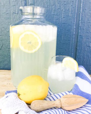 A glass pitcher of fresh squeed lemonade stand behind a glass of lemonade and a yellow lemon