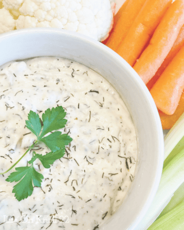 grandmas sour cream dill dip in a white bowl, topped with fresh parsley and surrounded by vegetables