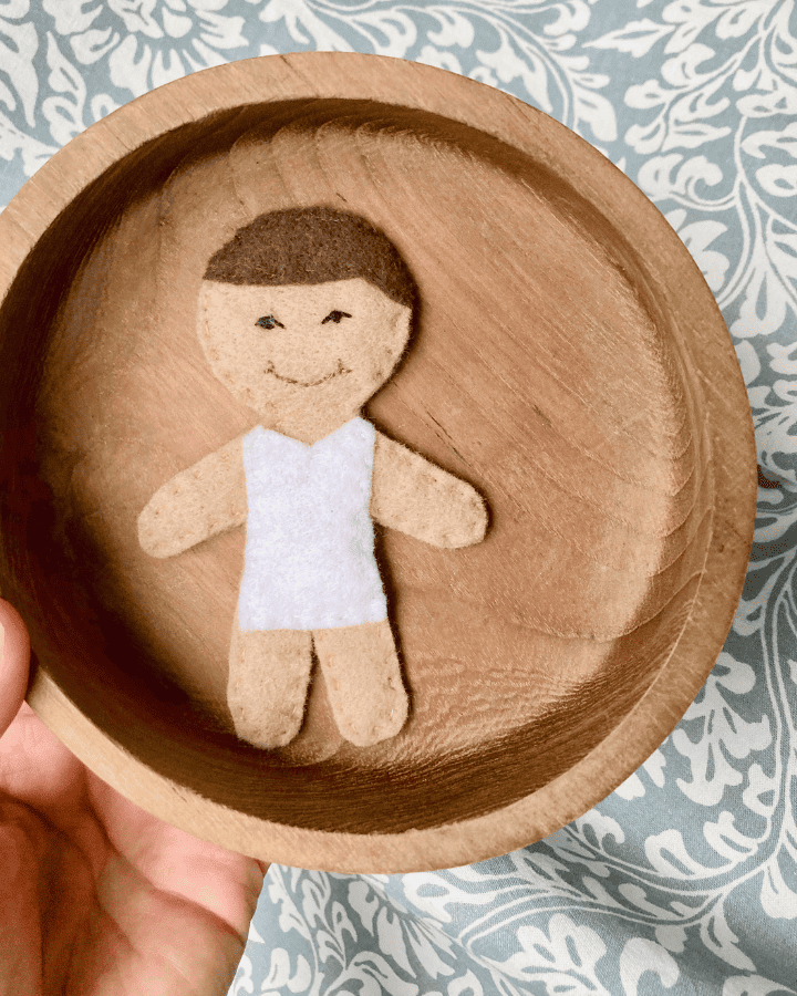 felt doll child wearing a white bodysuit laid in a wooden bowl 