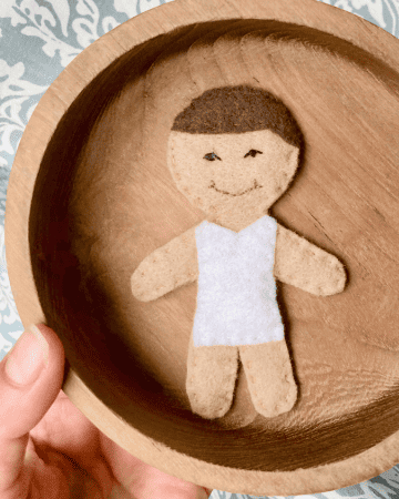 handmade felt doll child wearing a white bodysuit lay in a wooden bowl held over a blue and white tablecloth