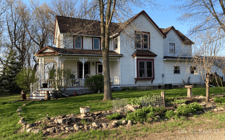 White Victorian farmhouse with burgundy red accents on windows and a brown roof.  flower bed in front with birdbath.